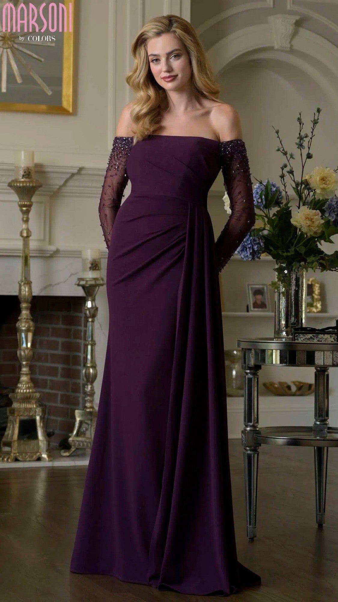 A woman stands indoors wearing the Marsoni M344 long formal dress in eggplant, featuring beaded details and sheer embellished sleeves, beside a table with a vase of flowers and decorative candles.